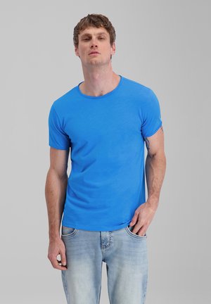 Man with short brown hair wearing a bright blue t-shirt and light blue jeans standing against a plain gray background.
