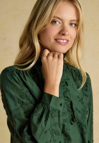 Dunkelgrüne Bluse mit floralem Stickerei, hohem Kragen, Rüschendetails und geknöpften Manschetten. Aus leichtem, strukturiertem Stoff gefertigt.