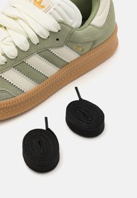 Zaļi zamša sporta apavi ar baltām ādas svītrām un zelta "Samba" logo. Blakus apavam novietoti divi melni apaļi šņorēm.