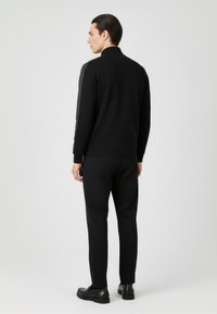 Czarna dresowa kurtka i spodnie wykonane z gładkiej tkaniny. Kurtka ma wysoki kołnierz i subtelny pasek na rękawach. Nosi się czarne buty.