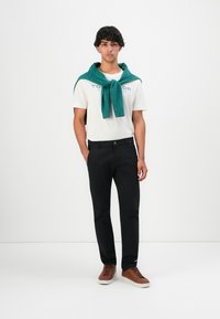 Ung mand står iført en hvid grafisk t-shirt, sorte bukser, brune sneakers og en grøn sweater bundet om sine skuldre foran en hvid baggrund.