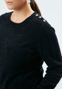 Sort fluffy sweater med rund halsudskæring og tre dekorative perleknapper på skulderen. Blød tekstur, tætsiddende design.