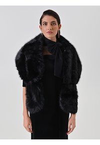 Châle en fausse fourrure noire avec une texture épaisse, col large et manches volumineuses. Le mannequin porte une robe noire et une cravate en tissu noire, design minimaliste.