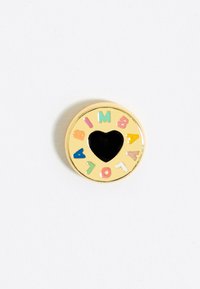 Pin circular en tono dorado con un centro en forma de corazón negro. El texto que lo rodea está en colores pastel y dice "BIMBA Y LOLA". Textura suave.