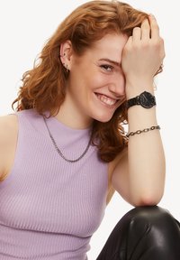 Schwarze Uhr mit rundem Zifferblatt und strukturiertem Armband, kombiniert mit einer silbernen Kettenhalskette, am Handgelenk getragen. Gestreiftes lilafarbenes Oberteil und schwarze Hose.