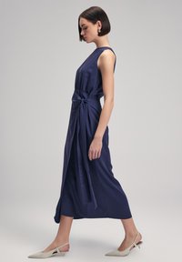 Navy blue sleeveless dress with a knotted waist detail, flowing fabric, and a midi length. White heels complement the outfit.