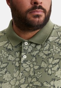 Grünes Polo-Shirt mit floralem Druck in Schwarz. Baumwollmischung mit klassischem Kragen und Knopfleiste mit drei Knöpfen. Strukturierte Oberfläche.