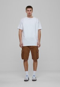 Hvit bomulls t-skjorte, løs passform. Brune cargo shorts med sidelommer. Modellen har på seg hvite sokker og grå joggesko med svarte detaljer.