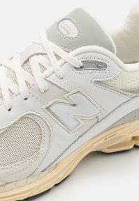 Gros plan d'une chaussure de course New Balance blanche et grise montrant le mesh, le daim, le logo, les lacets et une semelle beige avec détails d'amorti.