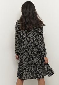 Zwarte jurk met een draaiend wit patroon, lange mouwen, een uitlopende zoom en een gestructureerde stof.