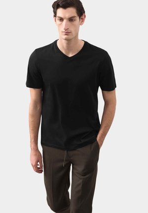 Jeune homme portant un t-shirt noir à col en V et manches courtes avec un pantalon marron foncé, debout, une main dans la poche, sur un fond clair.