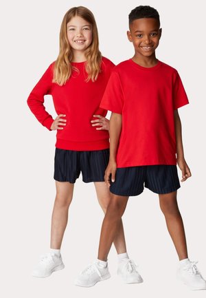 Un sweat-shirt rouge à manches longues, associé à un short plissé sombre. Modèle portant des baskets blanches. Vêtements pour enfants exposés ensemble.