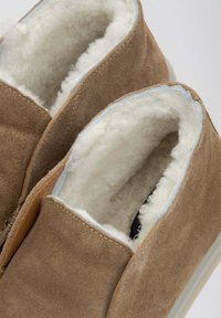 Brązowe zamszowe kapcie z miękką białą wyściółką. Zaokrąglony otwór, miękka faktura oraz jasnoniebieskie wykończenie na krawędzi. Brak widocznych elementów metalowych.