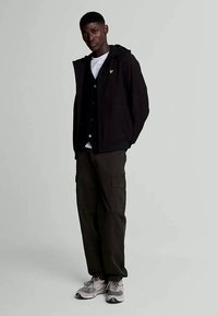 Black zip-up jacket with a hood, white T-shirt, black button-up vest, and olive cargo pants. Gray sneakers with white accents and logo.