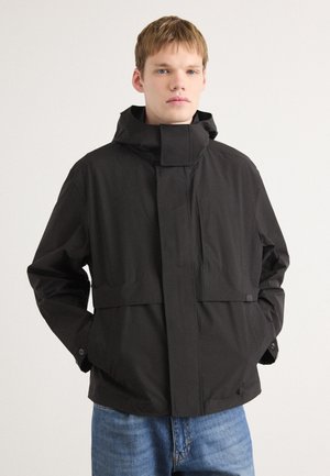 Giacca nera impermeabile con colletto alto, tasche anteriori con pattina e polsini regolabili. Caratterizzata da un design minimalista e da un tessuto leggero.