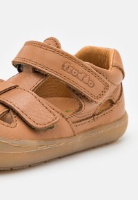 Brown leather sandal with velcro strap, open design, stitched accents, and a rubber sole. Features cutouts for ventilation.