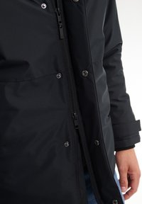 Giacca nera impermeabile con zip frontale e bottoni a pressione, realizzata in tessuto texture e con design aderente attorno ai polsi e all'orlo.