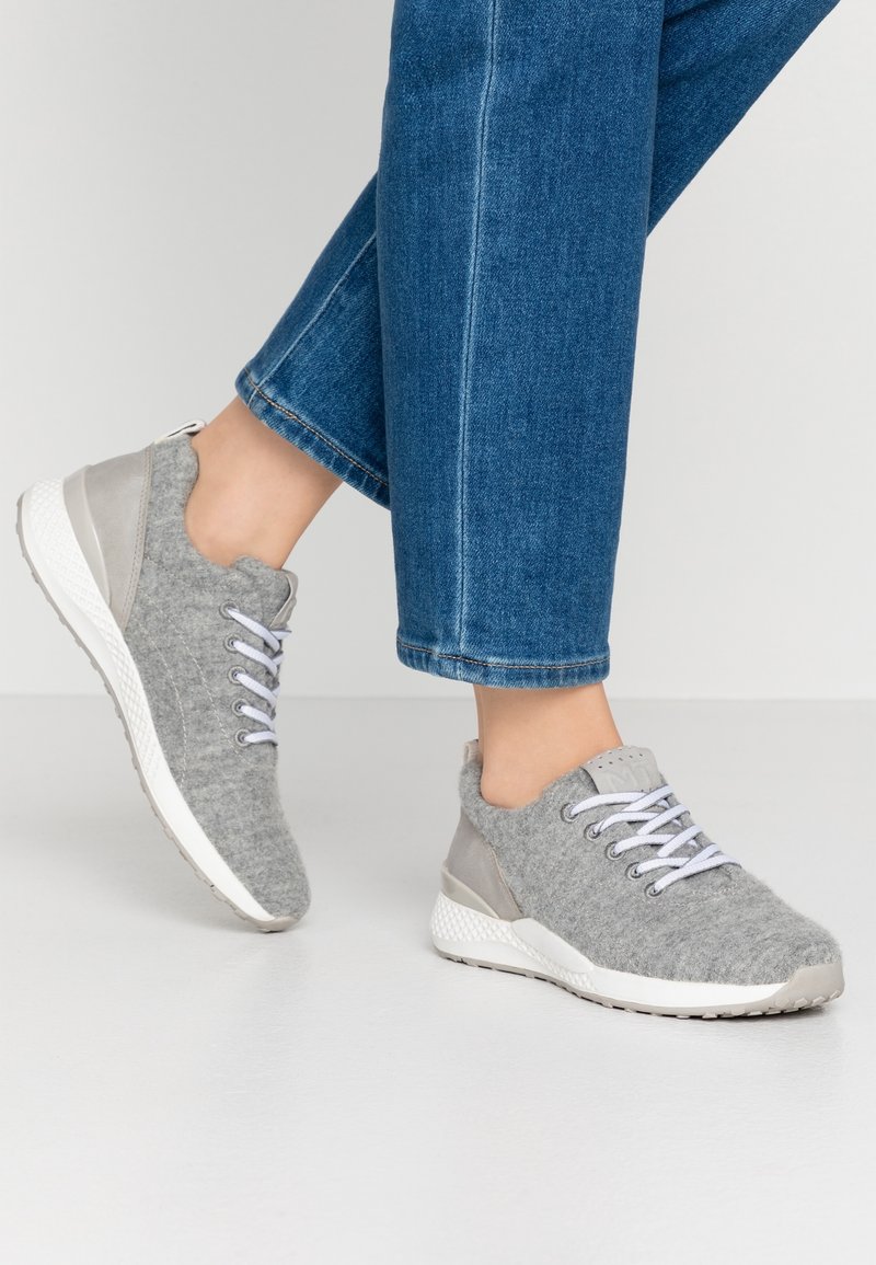 Pieds dans des sneakers en feutre gris avec des semelles et des lacets blancs, associés à un jean en denim bleu, sur un fond uni.