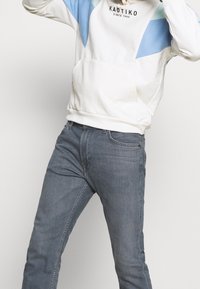 Vit hoodie med blå geometriska mönster och en känguruficka, kombinerad med mörkgrå denimjeans med klassisk passform.