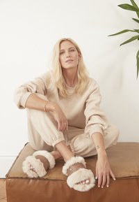 Beige loungewear-set med avslappnad passform, med elastiska ärmslut. Beige pälstofflor med bruna remmar och mjuk textur. Enkel träbakgrund.