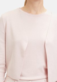 Světle růžový kardigan s žebrovanou texturou, otevřený vpředu, nošený přes odpovídající vršek. Hladká látka s bezešvými detaily.