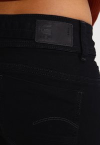 Jeans en denim noir avec une finition texturée, dotés d'une poche arrière cousue et d'une étiquette en cuir sur la ceinture.