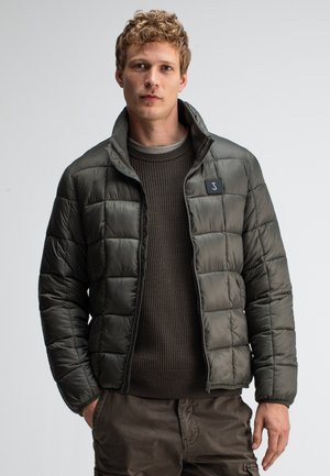 Olivgrüne Steppjacke mit einem gesteppten Design, Reißverschluss und Stehkragen. Trägt man über einem strukturierten braunen Pullover mit einem Logopatch.