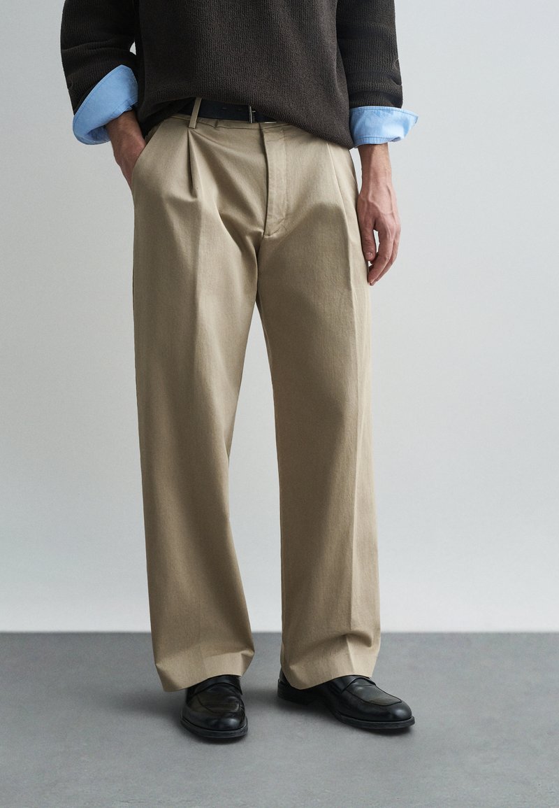 Šviesiai rudos klostuotos kelnės, pagamintos iš minkštos medžiagos, su laisvu siluetu. Derinamos su tamsiai pilku megztiniu ir juodais loaferiais.