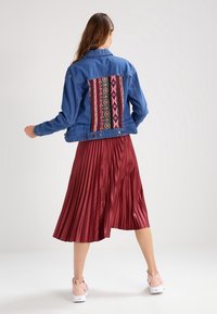 Giacca di denim blu con pannello posteriore ricamato colorato, abbinata a una gonna bordeaux plissettata. Indossata con sneaker rosa chiaro.
