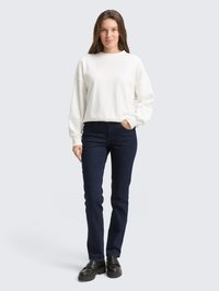 Mulher em pé, vestindo um sweatshirt branco de mangas longas, jeans azul escuro e sapatos mocassins pretos e robustos, com uma mão no bolso.