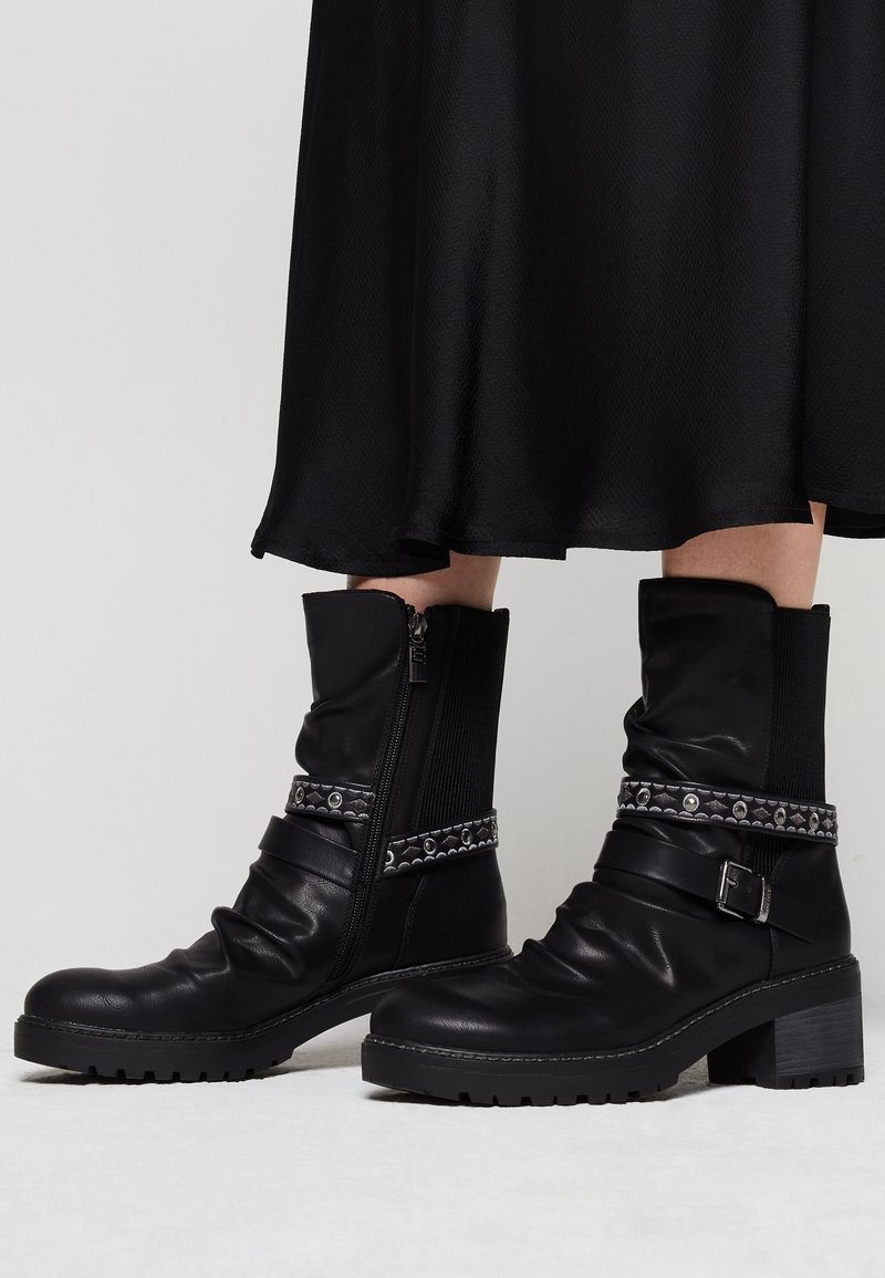 Bottes en cuir noires mi-mollet avec talons épais, sangles décoratives et fermetures éclair, portées avec une jupe mi-longue noire sur un fond gris.