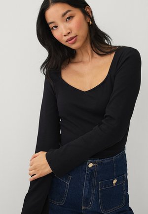 Kvinde iført sort langærmet V-hals top og højtaljet blå denimnederdel med forlommer, poserende med armene krydsede.