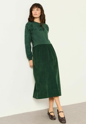 Donna con capelli scuri che indossa una maglia verde scuro a maniche lunghe con ricami floreali e una gonna midi abbinata, in piedi contro una parete chiara.