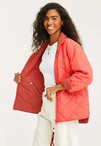 Kurtka z pikowanej tkaniny w kolorze koralowym, zapinana na zamek, z ribbowymi mankietami i wewnętrzną kieszenią; charakteryzuje się gładką fakturą i luźnym krojem.