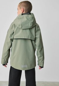 Lichtgroene waterdichte jas met capuchon, voorzien van een zwart logo label op de kraag en een gelaagd ontwerp. Zwarte broek maakt de outfit compleet.