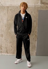 Fekete sportos melegítőfelső zipzárral és nadrággal, fehér Puma logóval. Fehér sneakers-szel van összeállítva, piros részletekkel egy texturált háttér előtt.
