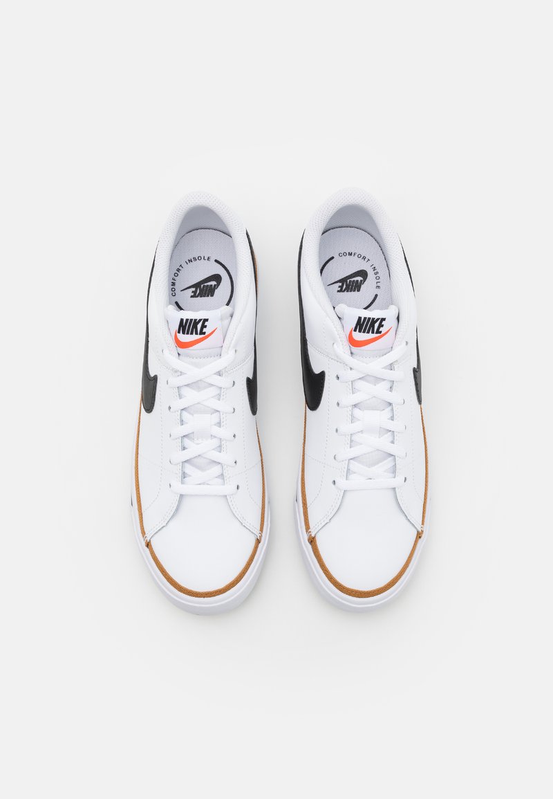 Zapatillas Nike blancas con un swoosh negro, acentos naranjas y suela de goma. Presentan una puntera redondeada y diseño con cordones. Plantilla cómoda.