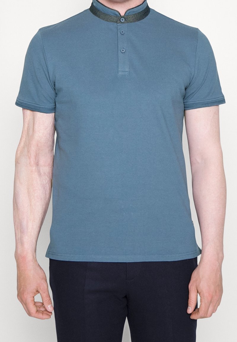Polo shirt teal à manches courtes, avec une patte de boutonnage et un col contrasté. Fabriqué en tissu doux avec une texture lisse.