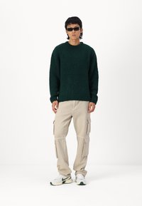 Pull en tricot texturé vert avec des poignets côtelés, associé à un pantalon cargo beige avec plusieurs poches, et des baskets blanches avec des accents verts.