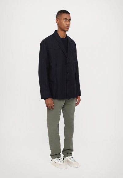 Navy gestreifter Blazer mit breitem Kragen, drei Knöpfen und zwei vorderen Taschen, kombiniert mit hellgrünen Hosen und weißen Sneakers.