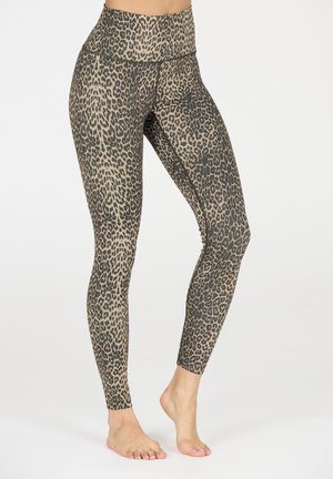 Vrouw in hoge taille, full-length leggings met luipaardprint, staand op blote voeten tegen een effen witte achtergrond.