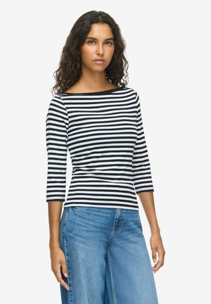 Un mannequin aux cheveux ondulés foncés porte un haut rayé noir et blanc à manches trois quarts et un jean bleu sur un fond blanc uni.