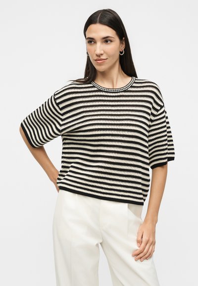 Γυναίκα με μακριά σκούρα μαλλιά που φοράει κοντομάνικο πουλόβερ με μαύρες και λευκές ρίγες και λευκό παντελόνι, στέκεται με το ένα χέρι στη μέση σε απλό φόντο.