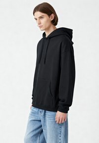 Sudadera de algodón negra con un amplio bolsillo frontal, capucha con cordón y puños acanalados. Llevada con jeans de mezclilla azul claro.