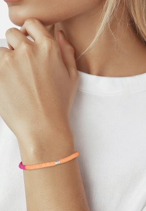 Primer plano de una muñeca con una pulsera de cuentas rosa y naranja llevada por una persona con piel clara y cabello rubio, vestida con una camisa blanca.