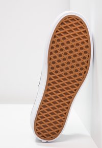 Semelle de chaussure avec motif de crampons en forme de gaufre marron et bord blanc, positionnée à la verticale contre une surface blanche.