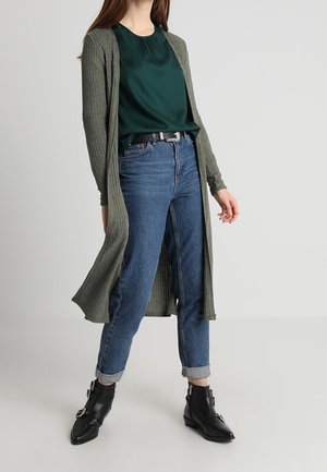 Cardigan lungo a coste di colore verde oliva sopra una blusa blu scuro, abbinato a jeans blu con risvolto e stivaletti neri alla caviglia con fibbie.