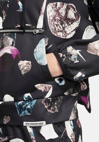 Sweat-shirt Nike en tissu noir avec des motifs de gemmes multicolores, une fermeture éclair à l'avant et une étiquette portant la mention "NIKE STANDARD ISSUE".