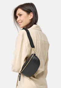 Zwarte leren halve maan tas met een gestructureerde afwerking, gouden ritssluiting en verstelbare schouderband, gedragen over een beige shirt.