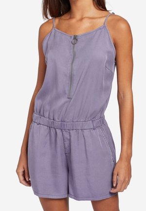 Kvinde iført ærmeløs lavendel romper med lynlås foran, elastisk talje og lommer, stående mod en hvid baggrund.